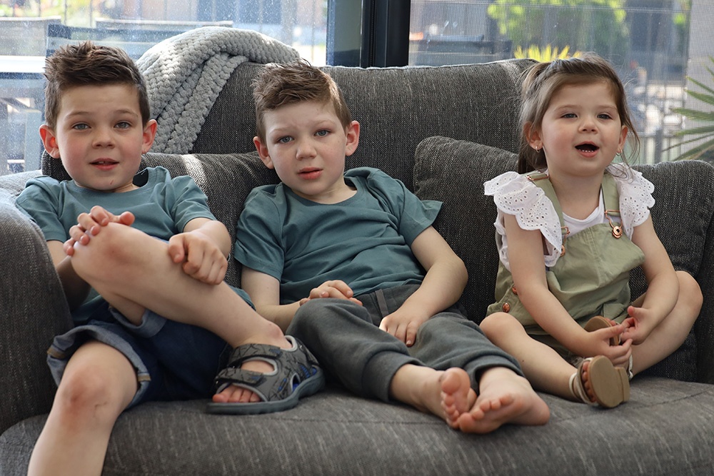 Two young brothers with their younger sister sitting on a couch smiling at the camera. 