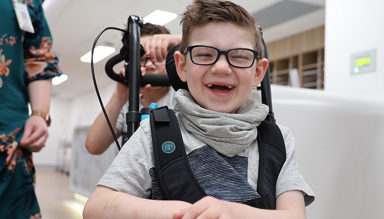 Young patient Declan in his wheelchair smiling at the camera being led by younger brother, Connor.