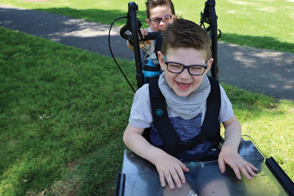 Young patient Declan laughing in a park being pushed in his wheelchair by younger brother, Connor.
