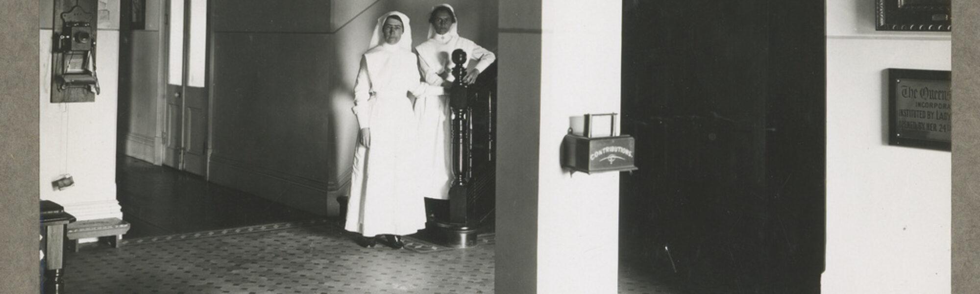 Two nurses stand at the bottom of the stairwell in the front lobby of Queen’s Home in Rose Park.