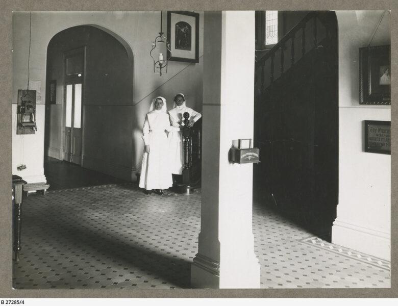 Two nurses stand at the bottom of the stairwell in the front lobby of Queen’s Home in Rose Park.